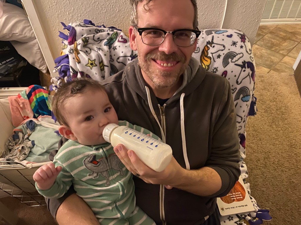 A smiling man holding a baby in a cozy setting, with the baby drinking from a bottle while wearing a green onesie.