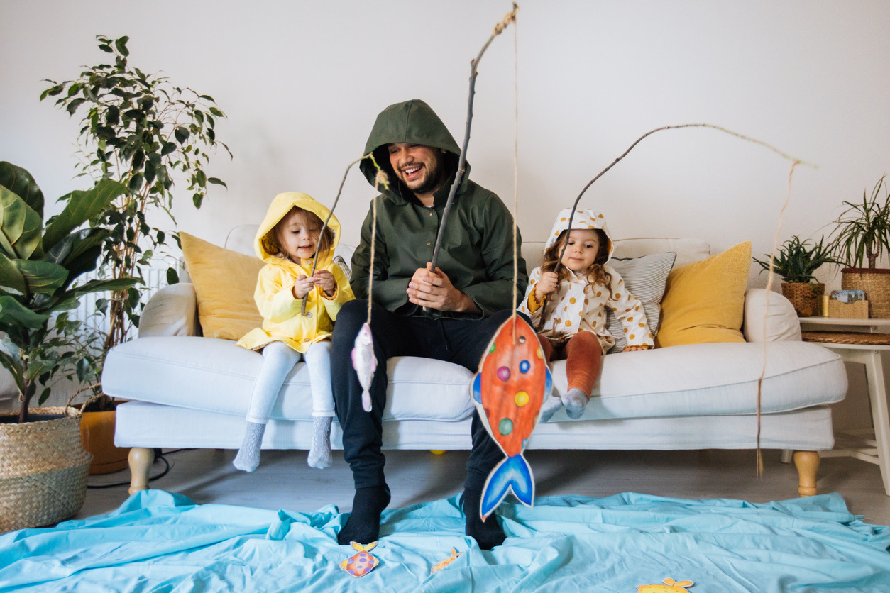 A man and two young children sit on a white couch wearing raincoats and holding makeshift fishing rods with paper fish attached. They're playing an imaginative indoor fishing game on a blue blanket spread on the floor, mimicking water. The setting is a cozy, plant-filled living room with a playful and joyful atmosphere.